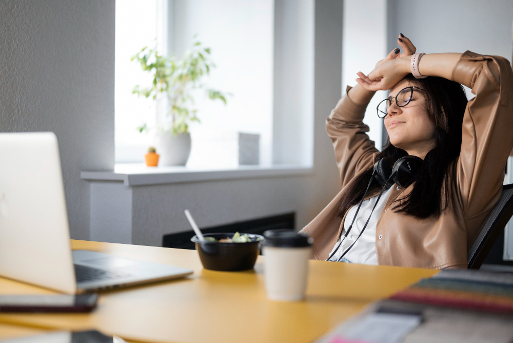 woman sitting relaxed at desk with headphones, food, and laptop