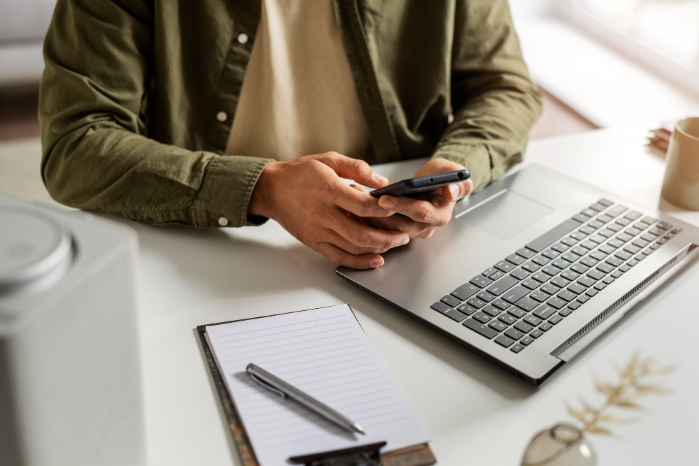 person using smartphone at desk with laptop and notebook